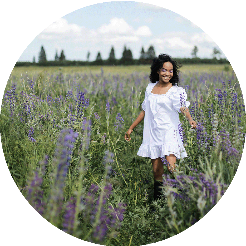 Smiling woman in a white dress standing in a field of purple flowers.